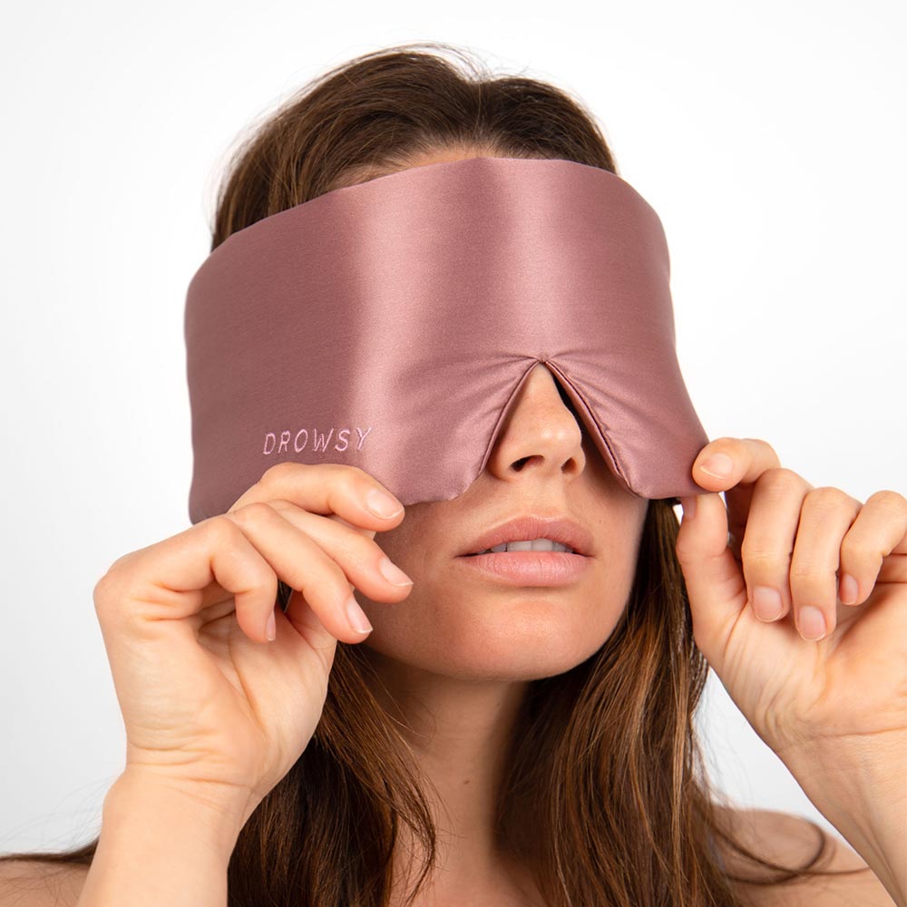 A woman adjusts a silky, rose-colored eye mask called Damask Rose over her eyes, holding the edges with both hands against a plain white background.