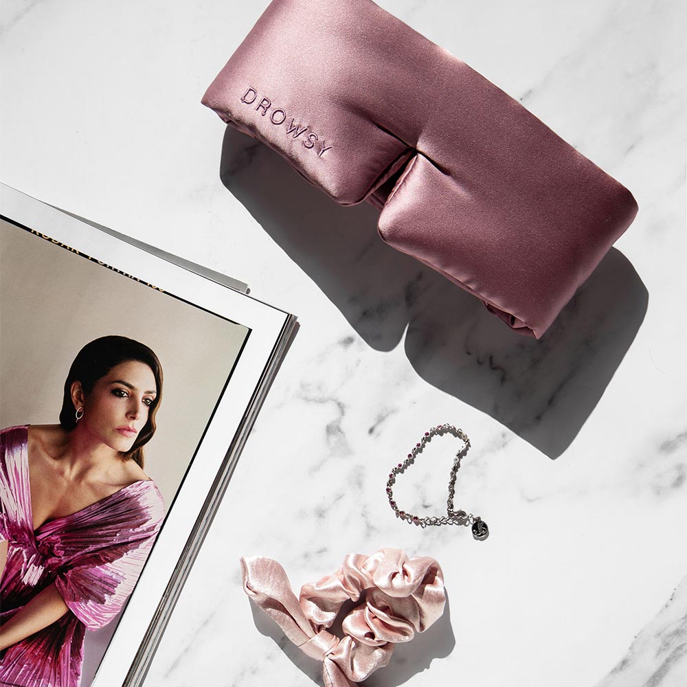 The Damask Rose pink silk eye mask, matching scrunchie, and silver bracelet rest on marble beside an open magazine featuring a woman in a pink dress.