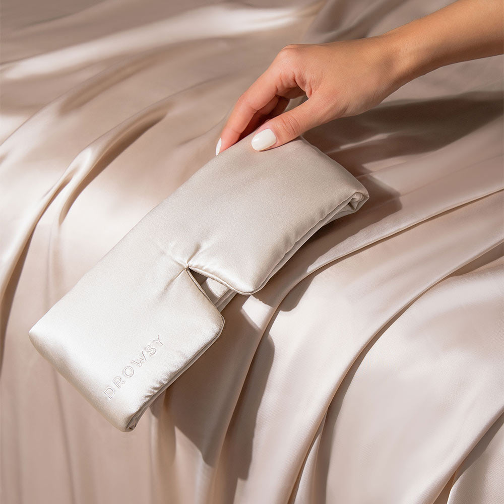 A hand holds the Dusty Gold silk eye mask over matching satin sheets, creating a smooth, luxurious look thats perfect for beauty sleep.