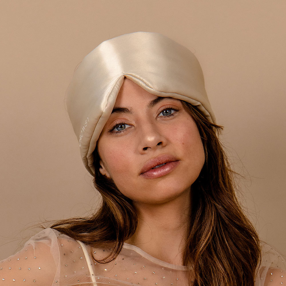 A woman with long brown hair wears the Dusty Gold satin turban and a sheer beaded top, posing before a beige background. She gazes calmly at the camera, capturing the tranquil elegance of beauty sleep.