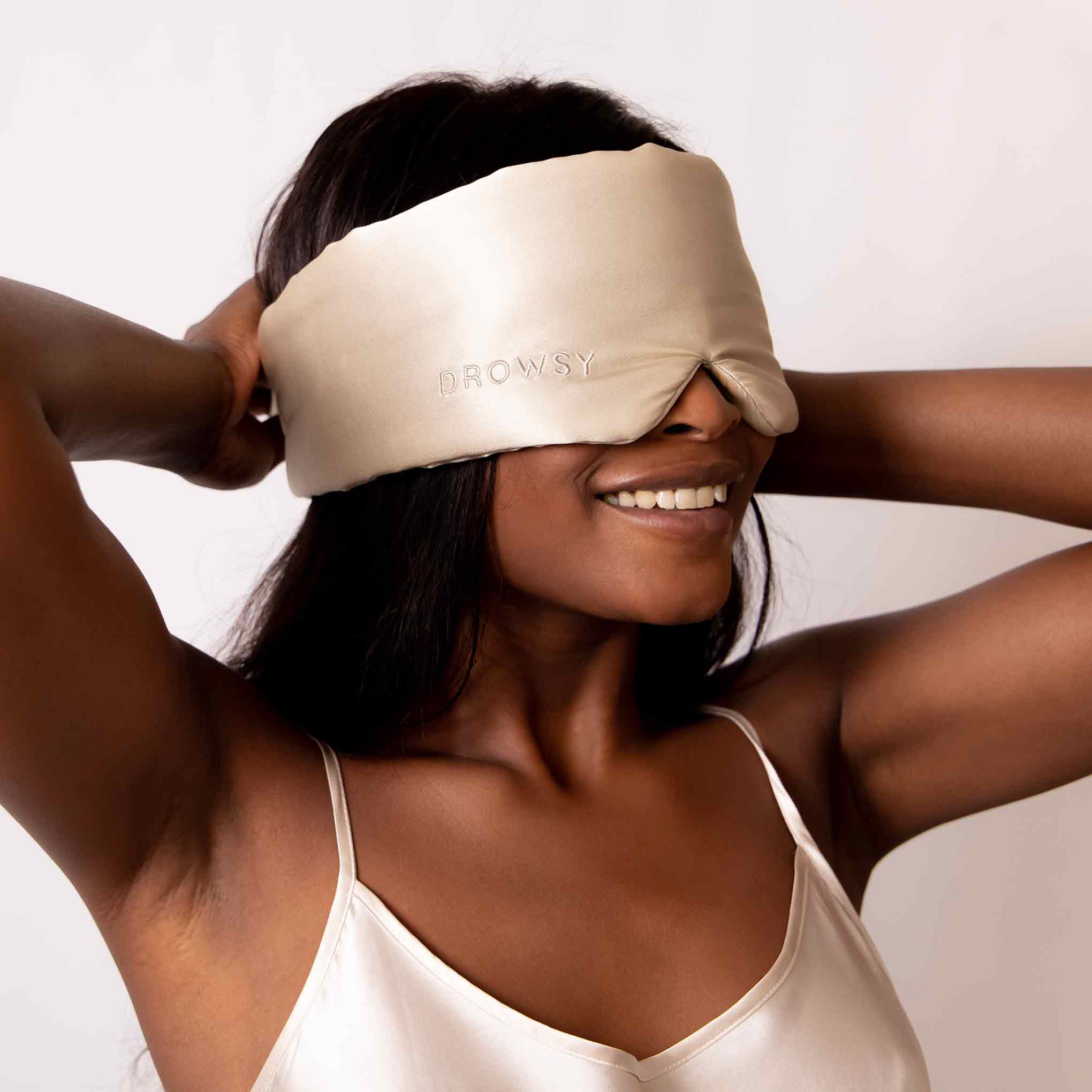 A woman wearing a beige silk sleep mask with the word DROWSY embroidered on it, smiling and adjusting the mask over her eyes. She is dressed in a matching satin camisole.