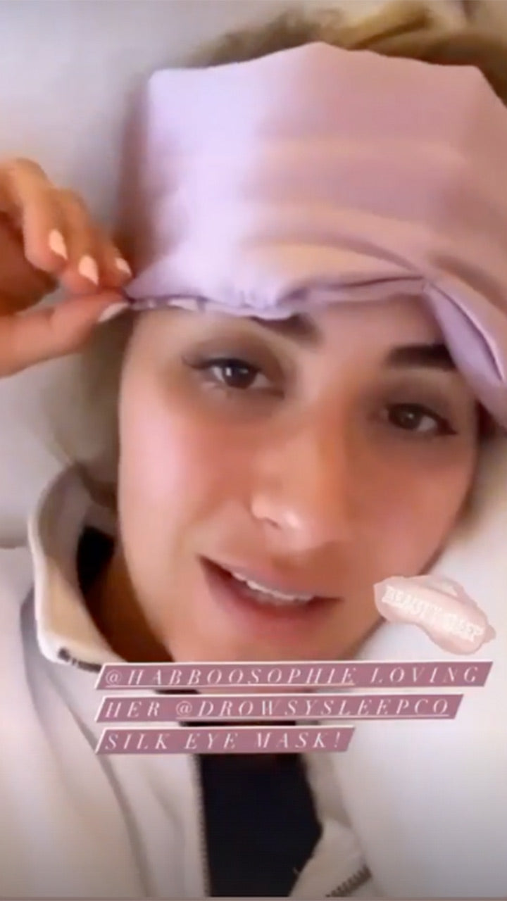 A woman lying down holds a light purple silk eye mask on her forehead. She has light skin and dark eyebrows, and is dressed in a light-colored top. Text on the image mentions loving a silk eye mask from a sleep company.