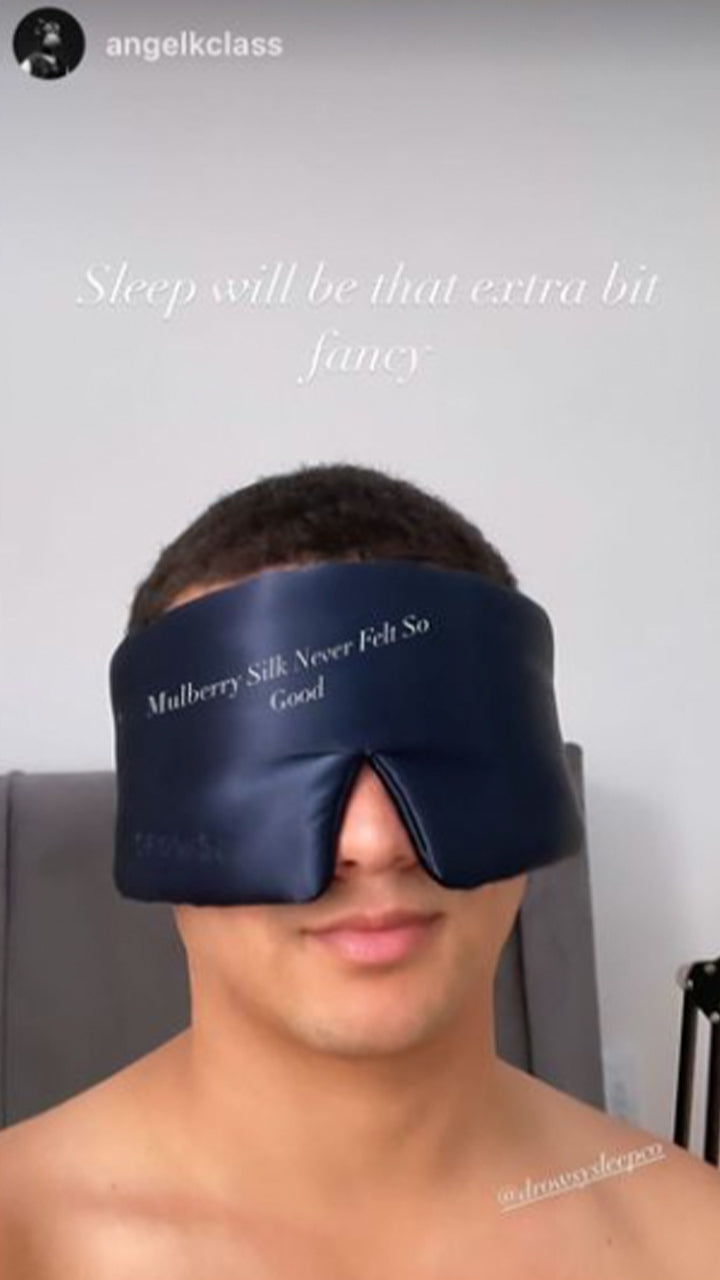 A person wearing a dark blue silk sleep mask that covers their eyes and part of their face. Text on the mask reads, Mulberry Silk Never Felt So Good. Text above says, Sleep will be that extra bit fancy.