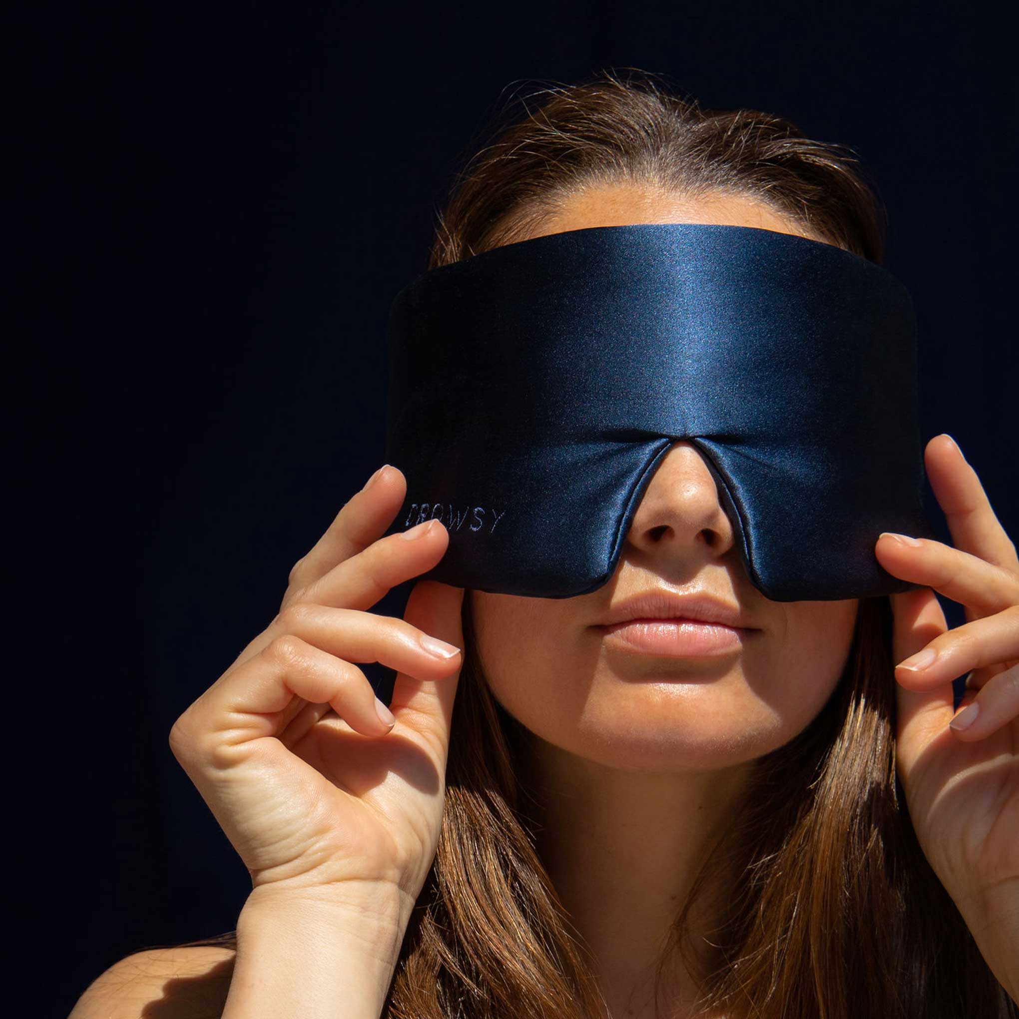 A woman with long brown hair holds a large, dark blue sleep mask over her eyes, adjusting it with both hands against a dark background.