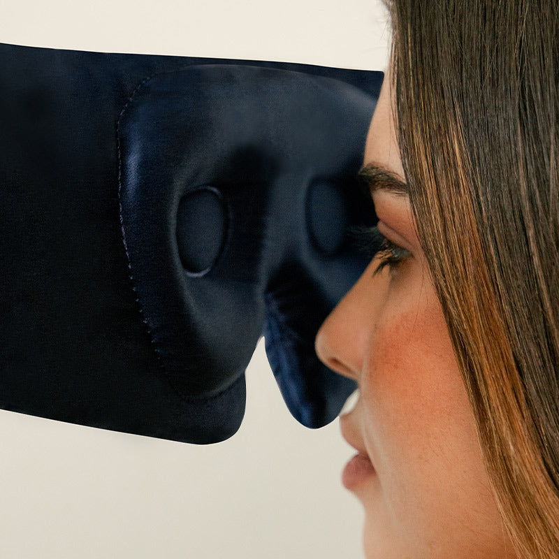 A woman with light brown hair wears a dark blue padded eye mask that covers her eyes and part of her nose, shown in a close-up side profile against a plain background.