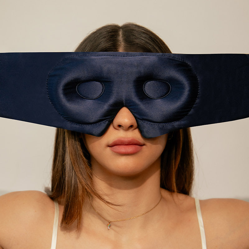 A woman with straight brown hair wears a wide, navy blue satin eye mask that covers her eyes and upper cheeks. She is facing forward against a plain, light background.