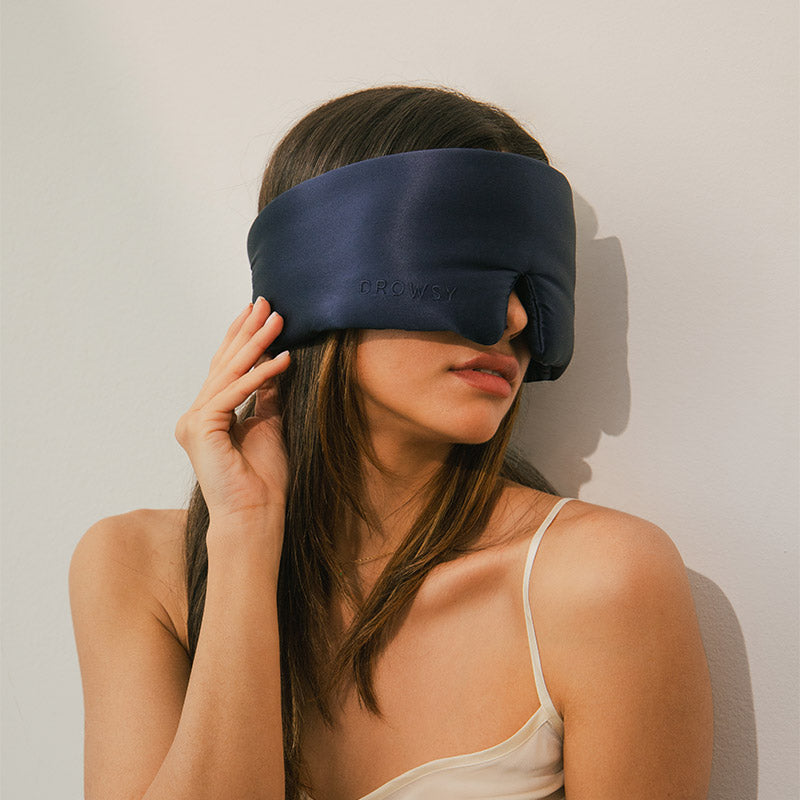 A woman with long brown hair wearing a navy blue sleep mask and a beige spaghetti strap top sits against a light-colored wall, gently touching the side of the mask.