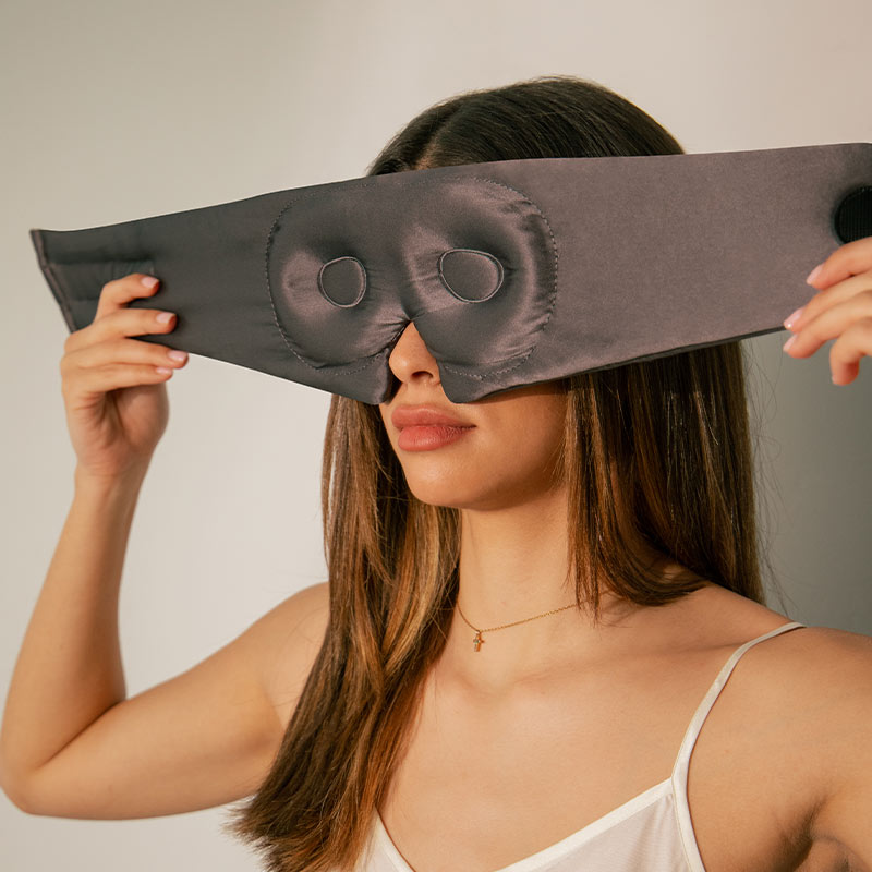 A woman wearing a white camisole puts on a black silk eye mask, holding it over her eyes with both hands. The mask has two round padded areas for the eyes.