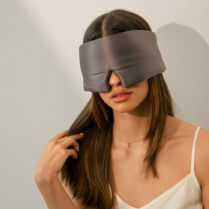 A woman with long brown hair wearing a gray, padded sleep mask sits against a light-colored wall. She touches her hair with one hand and wears a white spaghetti strap top.