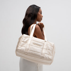 A woman with long dark hair carries the Drowsy Silk Weekender - Dusty Gold, a roomy quilted bag embroidered with DROWSY in gold, on her shoulder against a plain white background.