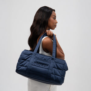 A woman with long dark hair in a white sleeveless outfit stands in profile, holding the spacious Drowsy Silk Weekender - Midnight Blue bag by Drowsy, featuring quilted navy silk with DROWSY embroidered on the side against a plain light background.