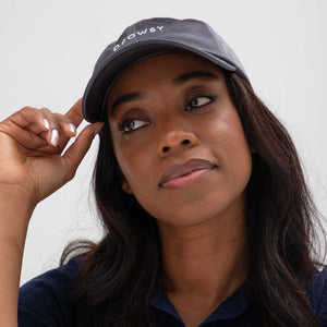 A woman with long dark hair wears the Drowsy Silk Cap - Moonlight Shadow, a stylish black cap offering sun protection and hair coverage as she looks up and to the side, her hand resting on the brim.