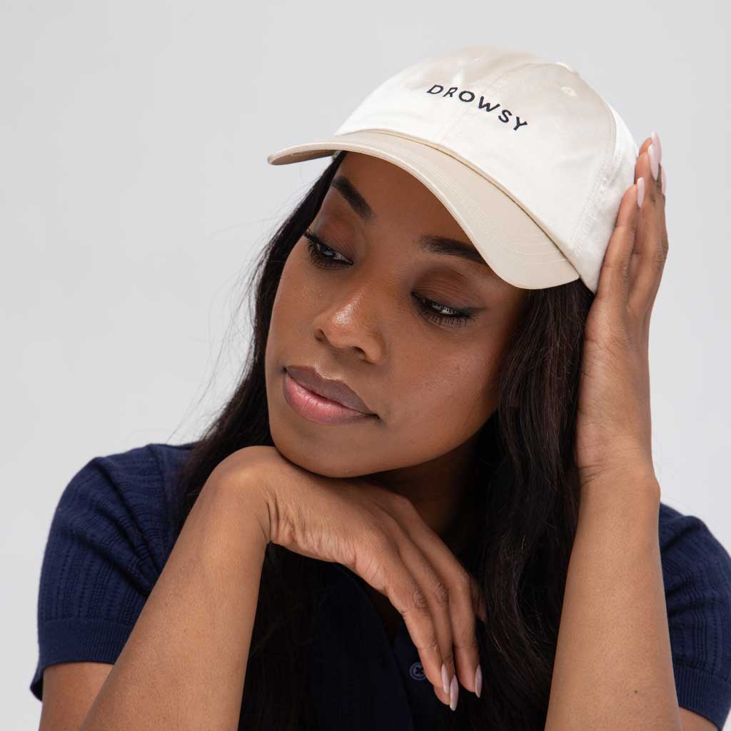 A woman wearing the Drowsy Silk Cap in Dusty Gold by Drowsy rests her head on her hand. The cap gently shields her face, complementing her long dark hair and navy blue top against a light background.