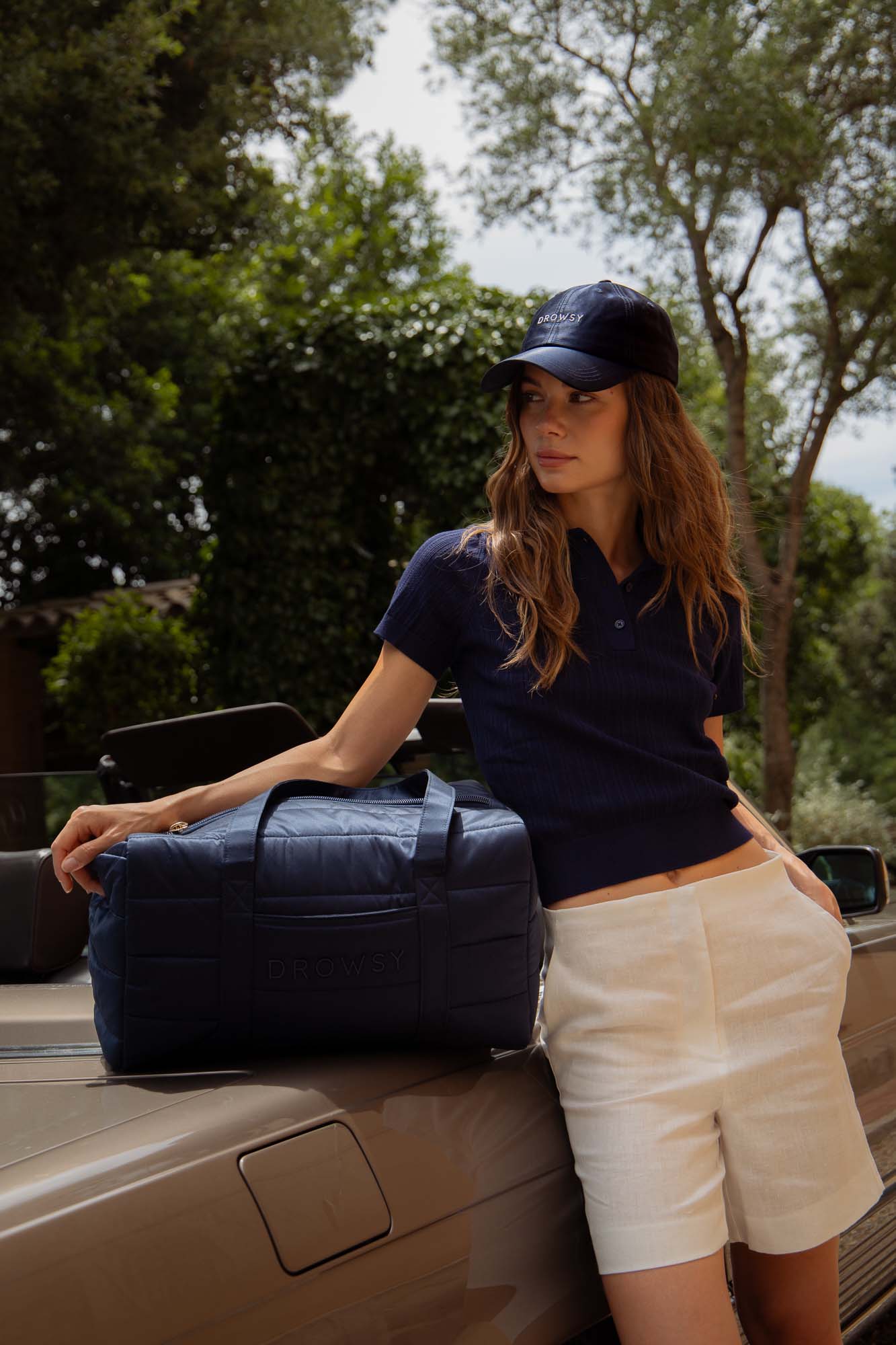 A woman wearing a navy cap, navy polo shirt, and white shorts leans against a convertible car. She rests her arm on a navy duffel bag labeled DROWSY. Trees and greenery are visible in the background.