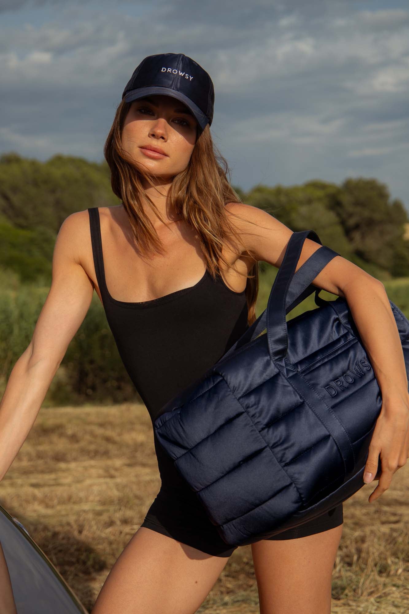 A woman wearing a black tank romper and a navy cap stands outdoors, holding a navy padded duffel bag embossed with “Drowsy.” Trees and grass are visible in the background under a cloudy sky.