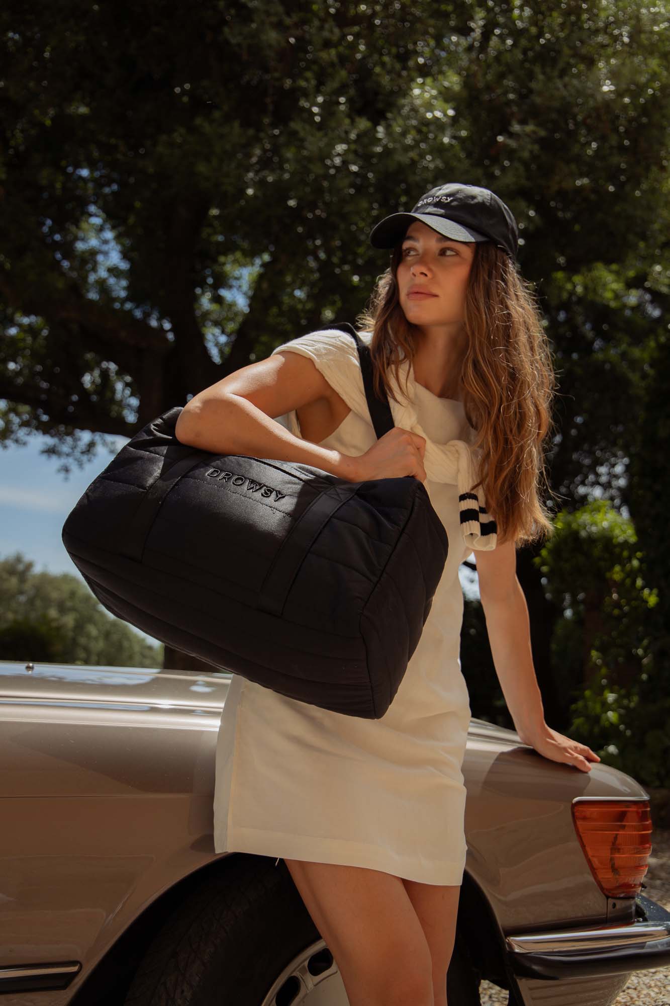 A woman wearing a black cap and white dress leans against a beige car, holding a large black duffel bag over her shoulder. She looks into the distance, with trees and blue sky in the background.