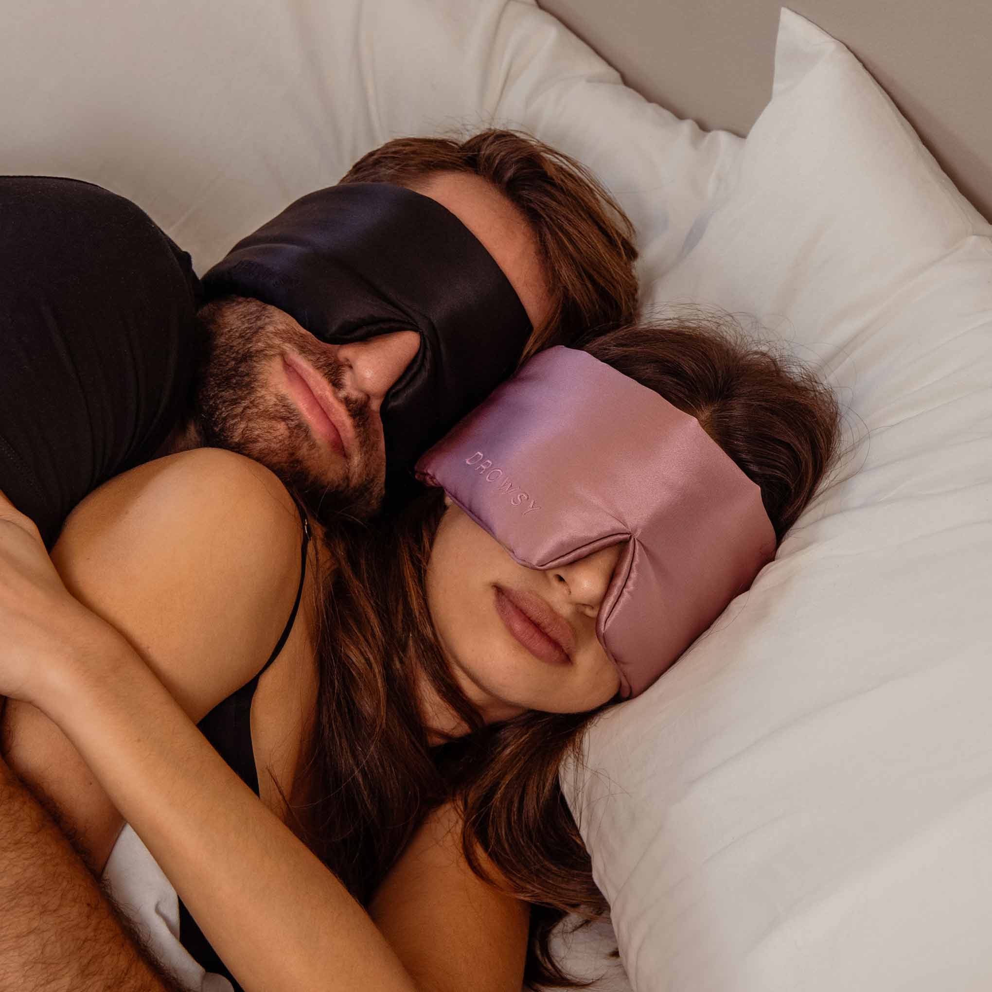 A couple snuggles in bed, each wearing a satin sleep mask from the Lovers Pack—one black, one pink—as they rest on white pillows.