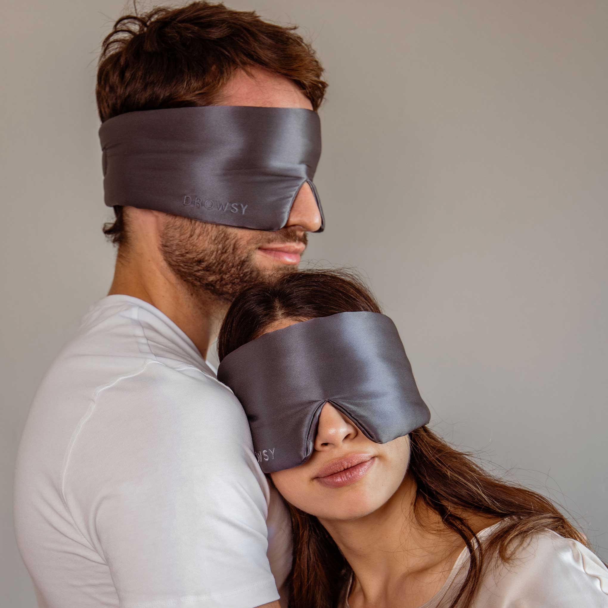 A man and woman wearing the Lovers Pack—large, dark gray silk sleep masks—stand closely together in white t-shirts against a plain background, with the woman resting her head on his chest.