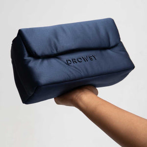 A hand holds up the Drowsy Silk Necessaire in Midnight Blue, a rectangular silk pouch featuring DROWSY embroidered in black letters—an elegant beauty accessory against a simple, light background.