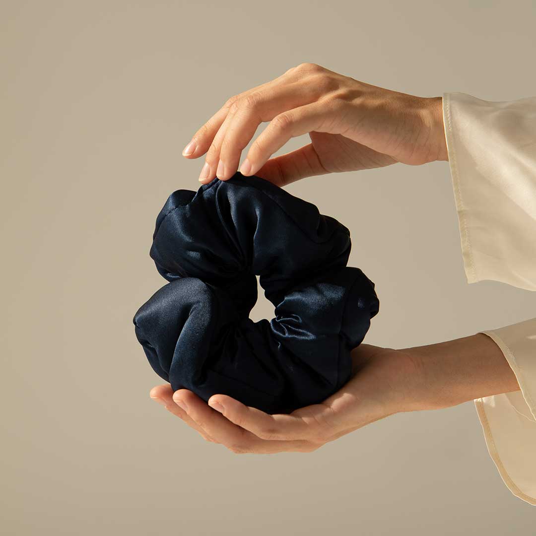 Two hands gently hold a large, dark blue satin scrunchie against a neutral background. The person wears a light-colored, long-sleeve garment.