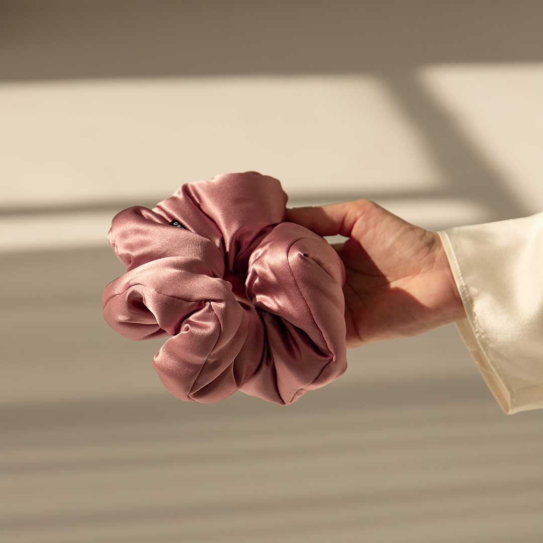 A hand in a white sleeve holds the Damask Rose - Padded Scrunchie against a softly lit, neutral background with gentle shadows.