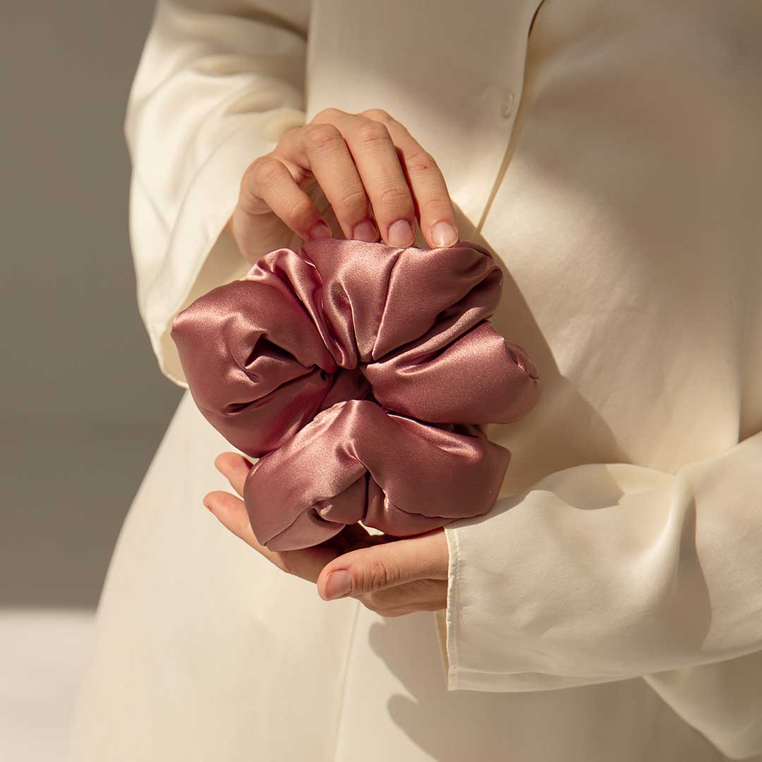 A person in a cream-colored satin shirt holds a large, shiny pink satin scrunchie with both hands, with soft natural light illuminating the scene.