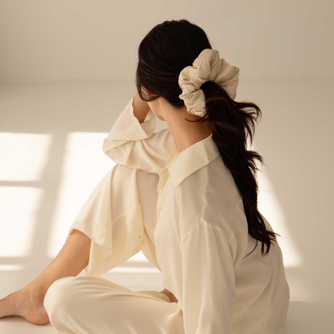 Woman with long dark hair tied in a loose ponytail with a large cream scrunchie, wearing light-colored pajamas, sits on the floor in soft, natural light, looking away from the camera.
