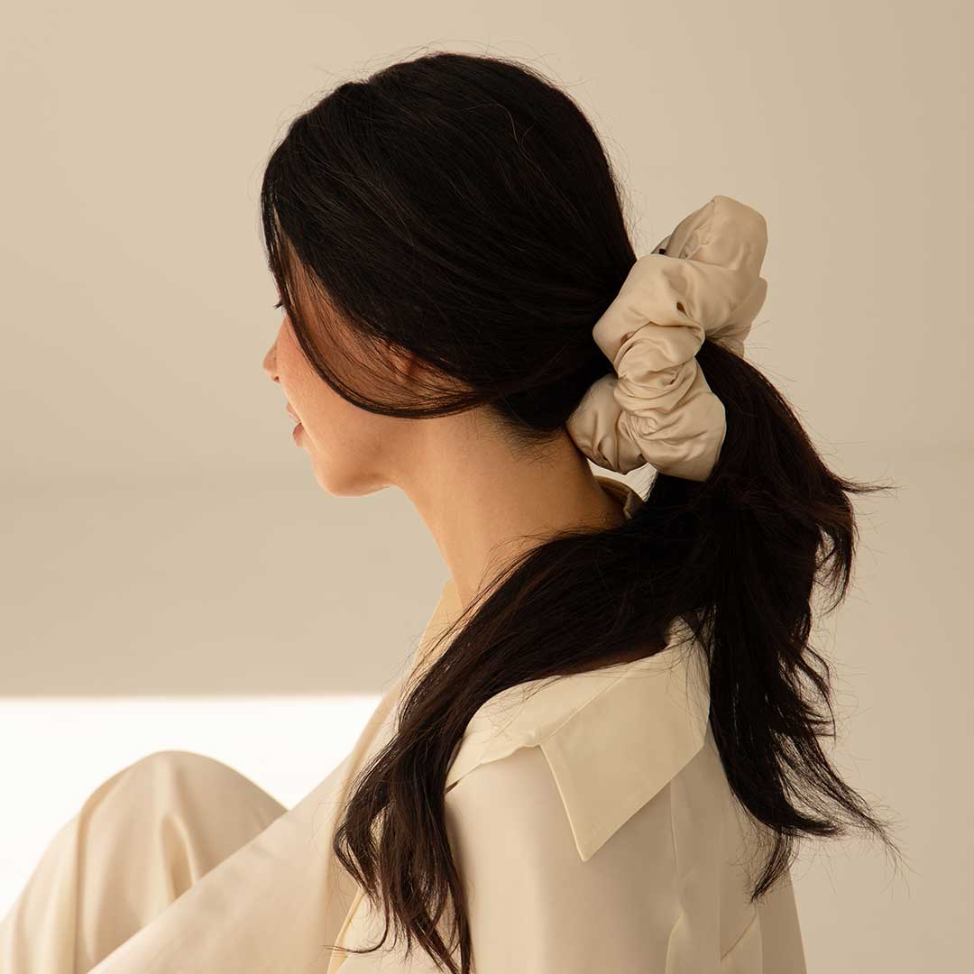 A woman with long dark hair in a low ponytail, secured with the Dusty Gold - Padded Scrunchie, sits sideways wearing a loose cream shirt against a neutral backdrop.