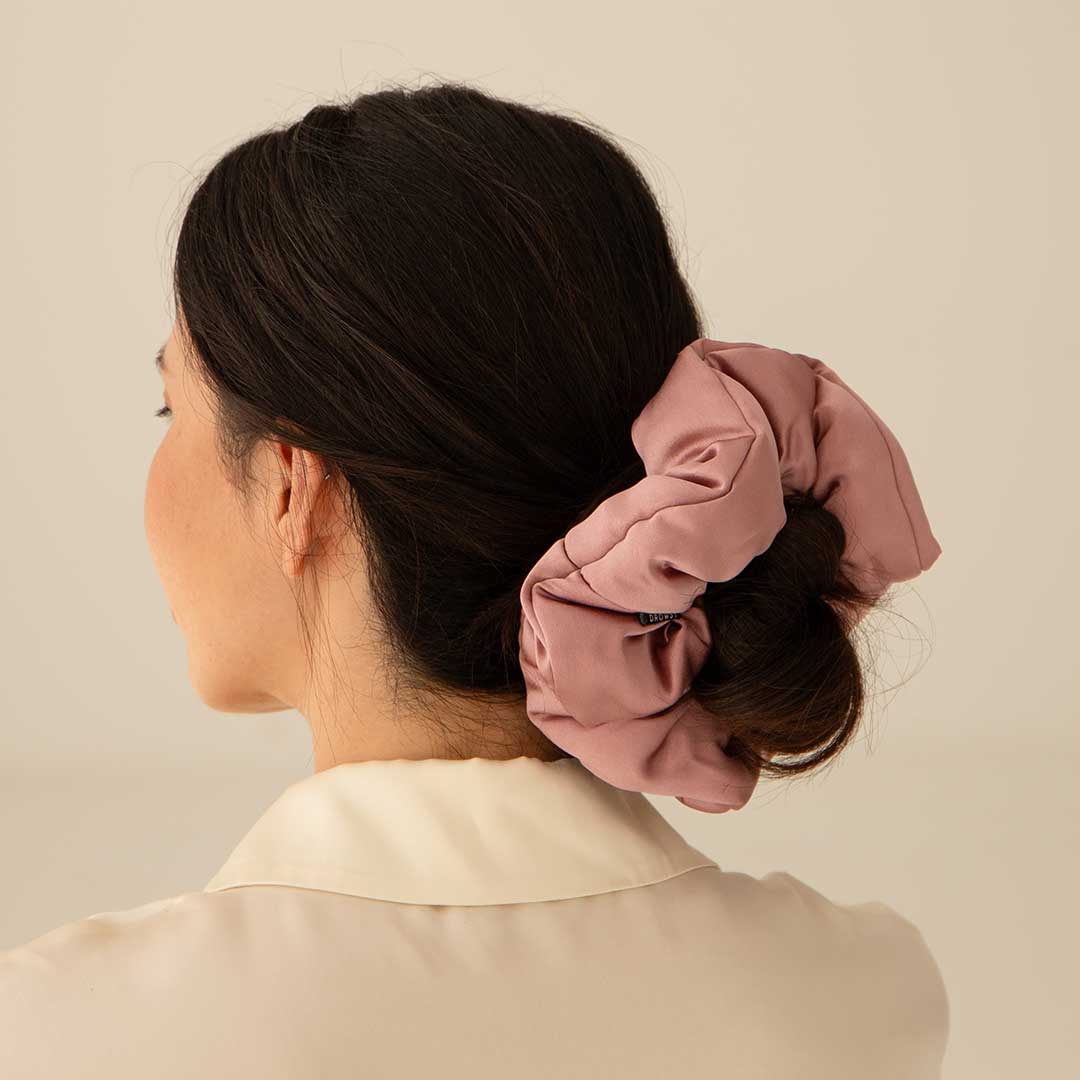 A woman with dark hair in a low bun wears the Damask Rose - Padded Scrunchie, oversized and light mauve, paired with a cream collared shirt. Shes seen from behind against a plain background.