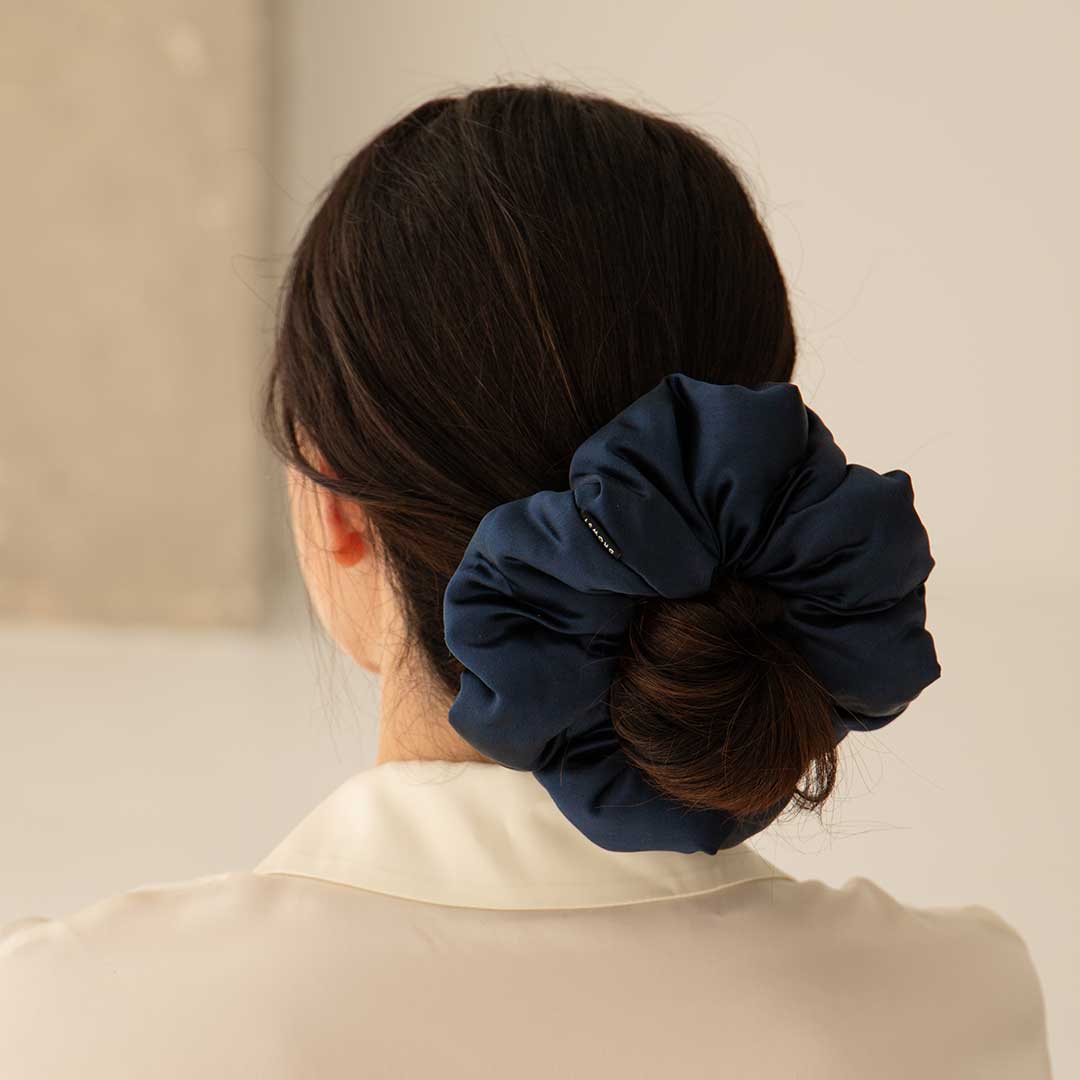A woman with dark hair in a low bun wears the Midnight Blue - Padded Scrunchie, adding volume to her style. Seen from behind in an off-white blouse, she stands out against a softly blurred background for a gentle effect.