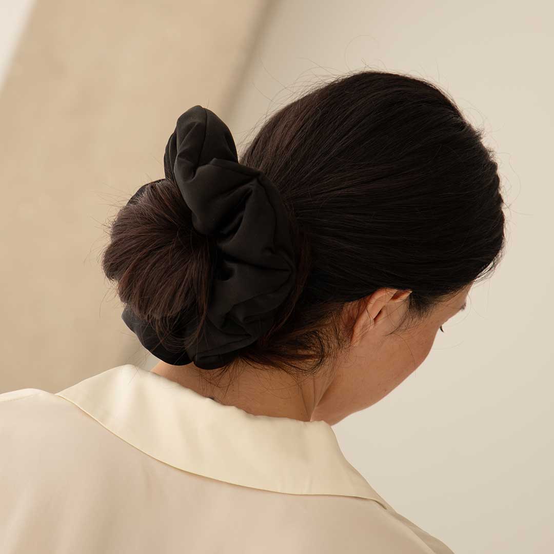 A woman with dark hair in a low bun, styled with the Black Jade - Padded Scrunchie, wears a cream collared shirt and is shown from behind against a neutral background.