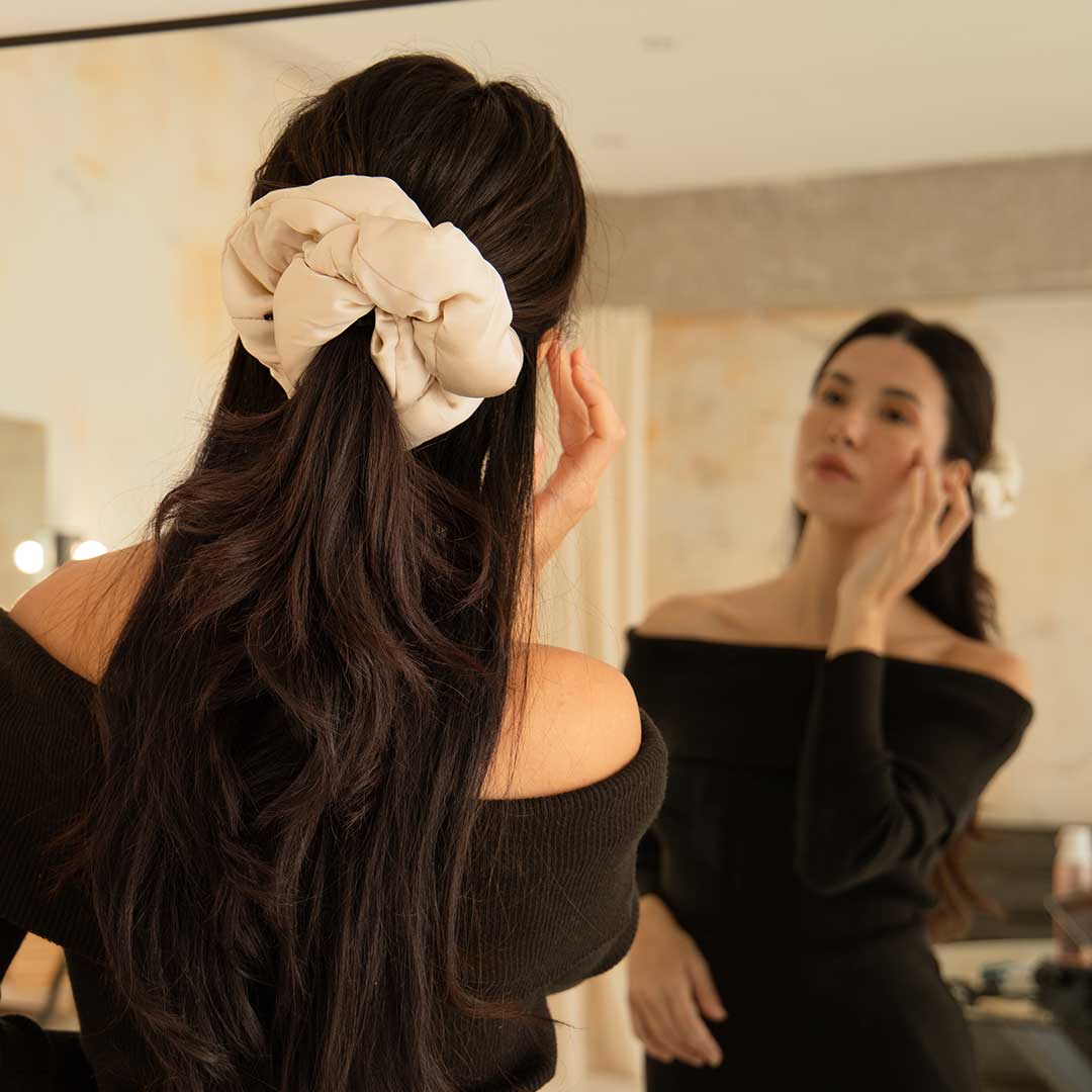 A woman with long dark hair, wearing an off-shoulder black top, adjusts her face in front of a mirror. Her hair is styled with the Dusty Gold - Padded Scrunchie. The background features a softly lit room.