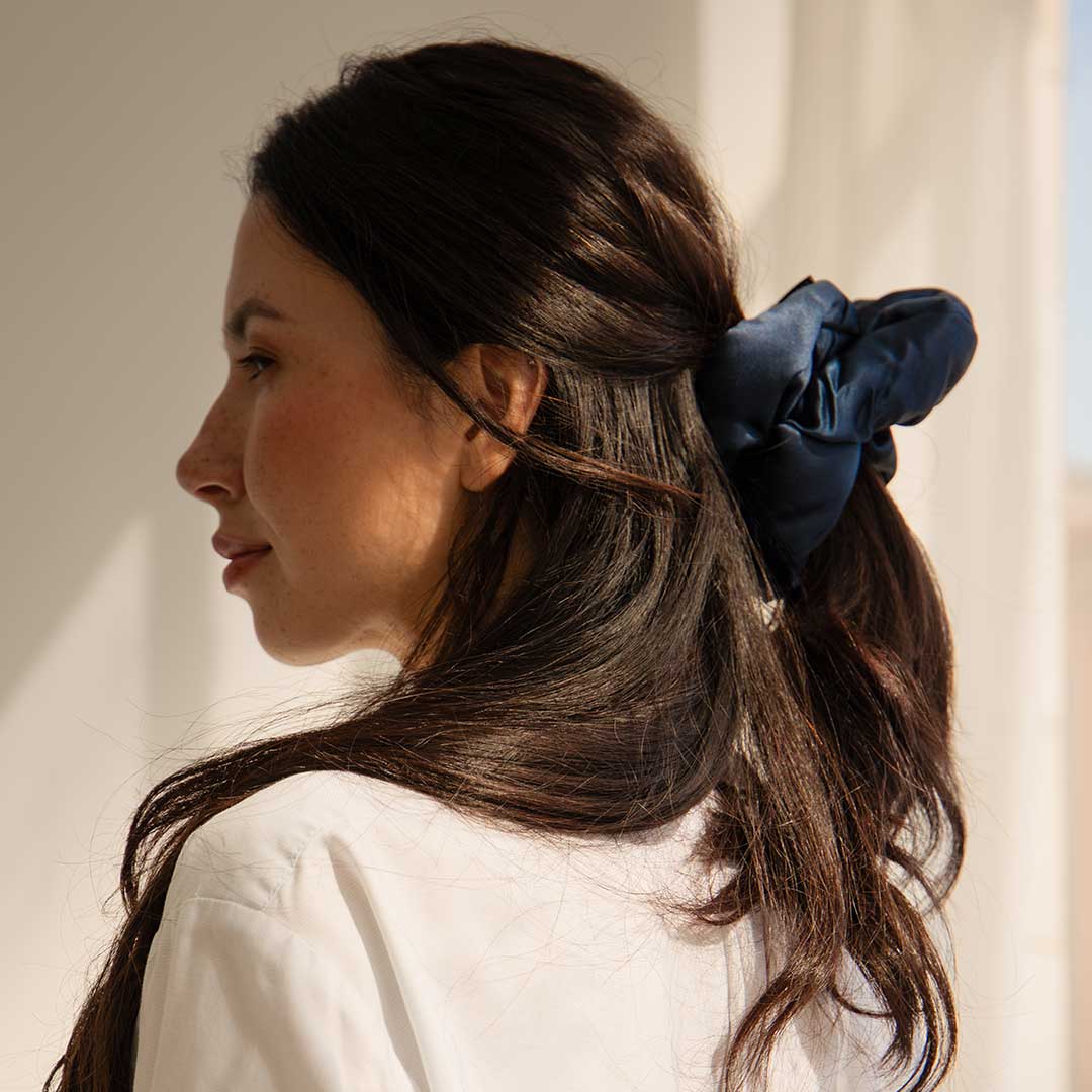 A woman with long dark hair, styled in a low ponytail with a large navy blue scrunchie, wears a white shirt and looks out towards soft natural light coming through a window.