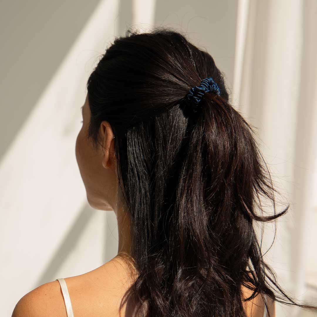 A woman with long, dark hair tied in a ponytail with a dark scrunchie, wearing a white strap top, is shown from behind in front of light-colored curtains with sunlight casting shadows.