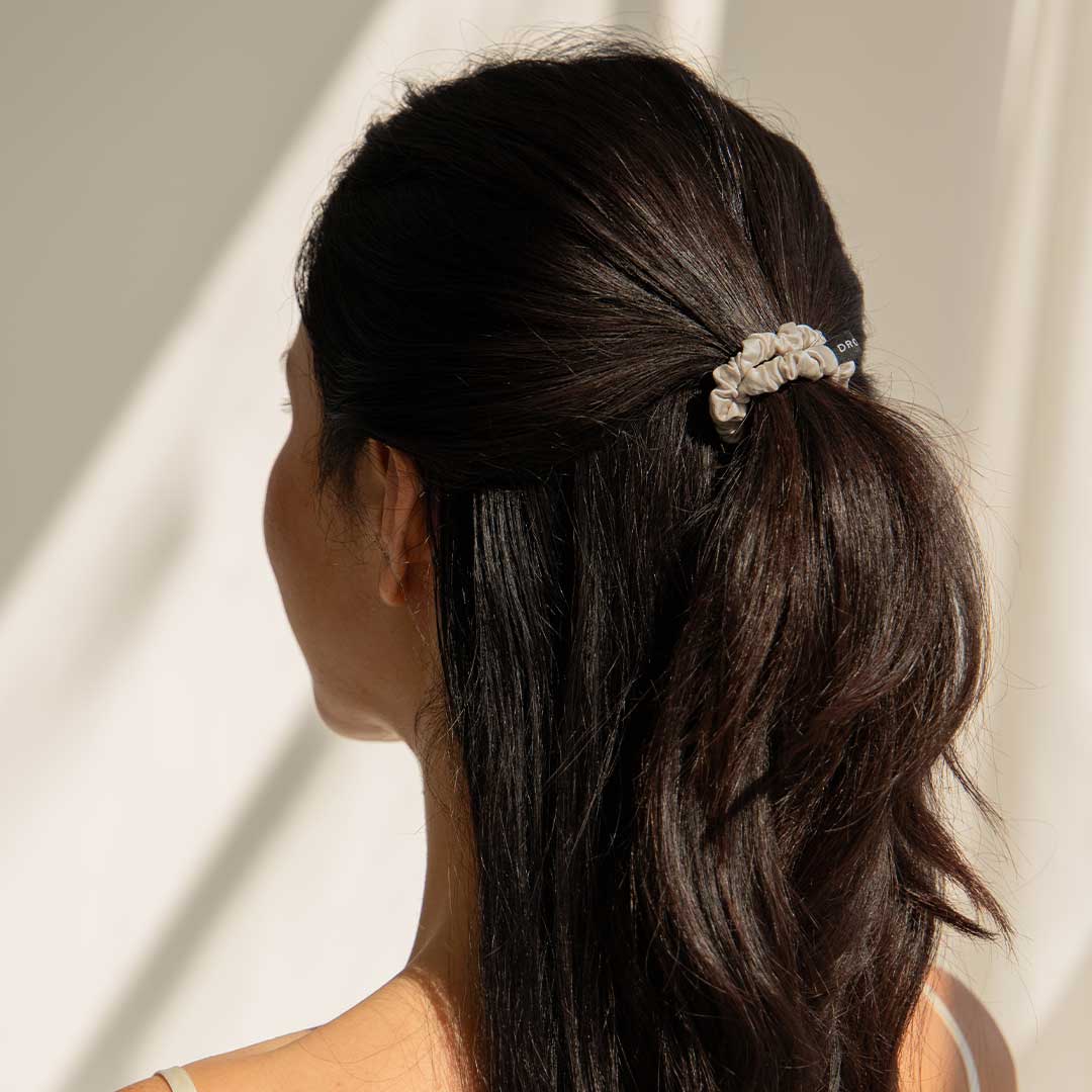 A woman with long, dark hair is seen from behind, wearing a white scrunchie to tie her hair in a loose ponytail. The background is softly lit with natural light.