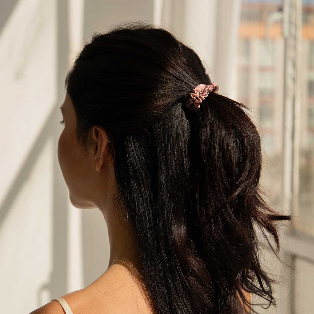 A woman with long dark hair, styled in a half-up ponytail with a pink scrunchie, stands near a sunlit window, facing away from the camera.