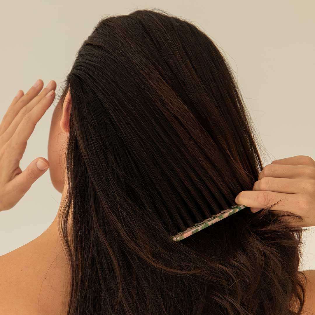 A person with long, dark hair is seen from behind, gently combing their hair with a patterned comb while raising their other hand near their head. The background is plain and neutral.