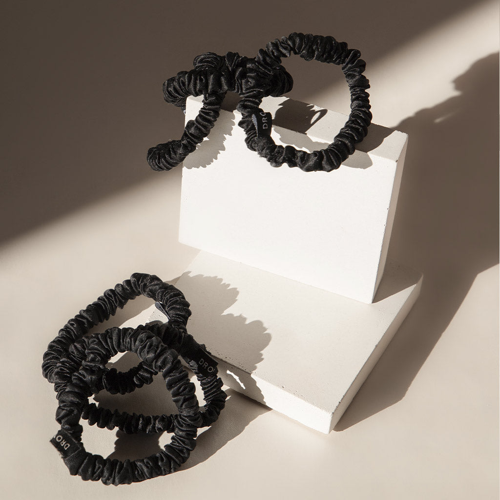 Three Skinny Scrunchie Set - Black Jade silk scrunchies are displayed on two white geometric blocks with striking shadows. Made from 22 momme silk, these black scrunchies have a softly shiny, gathered finish.