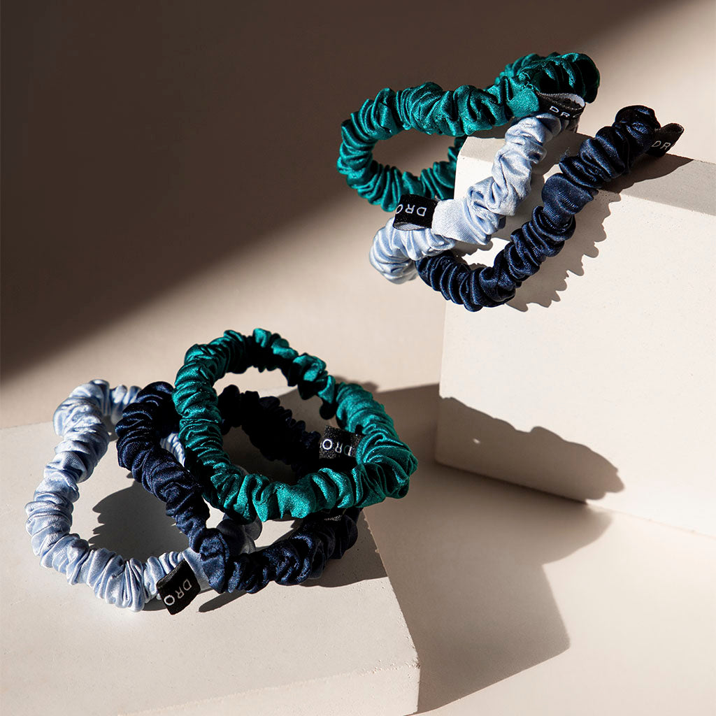 The Skinny Scrunchie Set - Blue & Green Shades features three silk scrunchies in green, light blue, and navy displayed on off-white blocks with sunlight casting soft shadows—ideal for gentle hair protection all day.