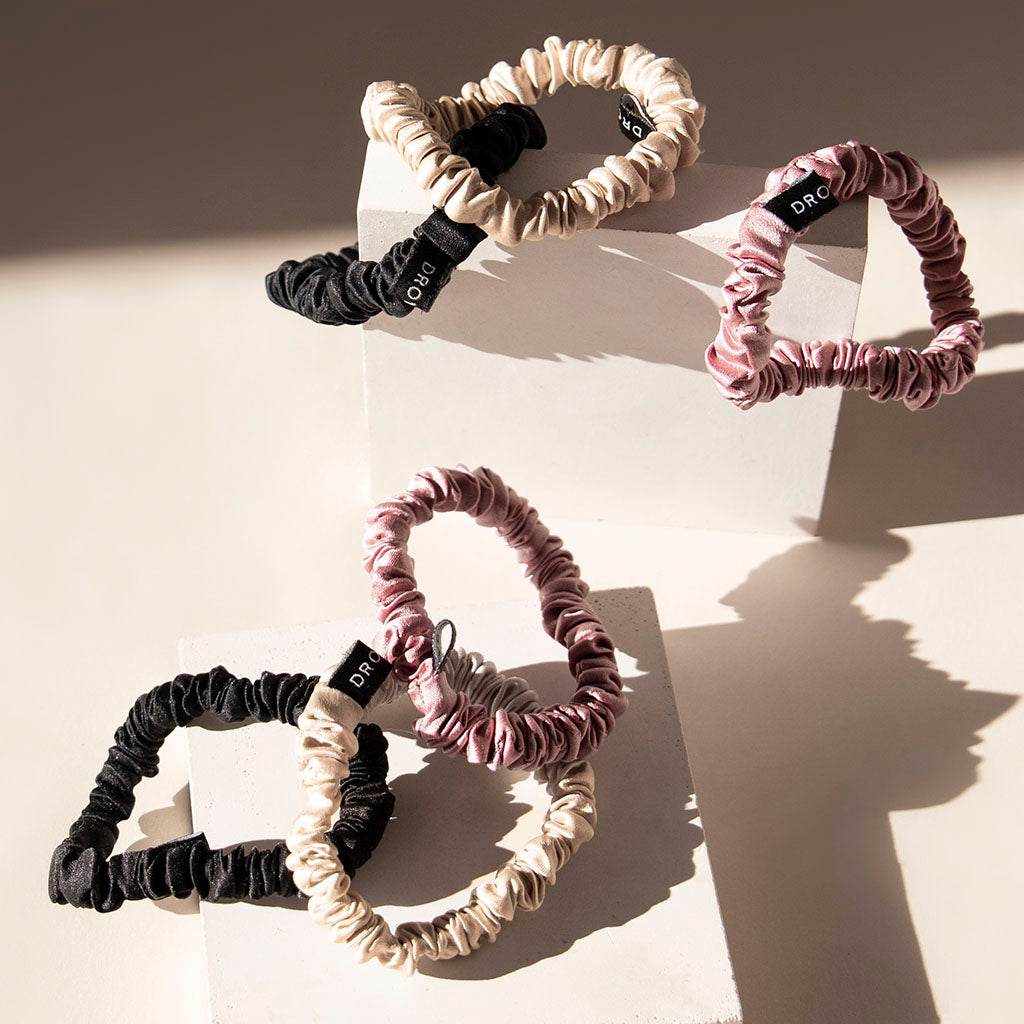 The Skinny Scrunchie Set—featuring ruched silk scrunchies in Rose, Dusty, and Black, each with a subtle black label—is displayed on white blocks, offering gentle hair protection under soft sunlight.