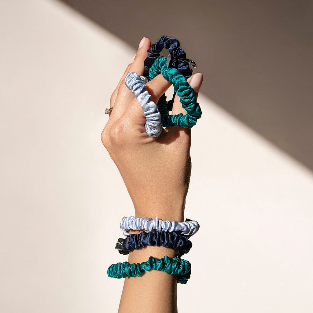 A hand displays the Skinny Scrunchie Set - Blue & Green Shades, featuring navy, light blue, and green silk scrunchies for gentle, crease-free hair protection. Extras are worn on the wrist. Cream background with visible shadow.