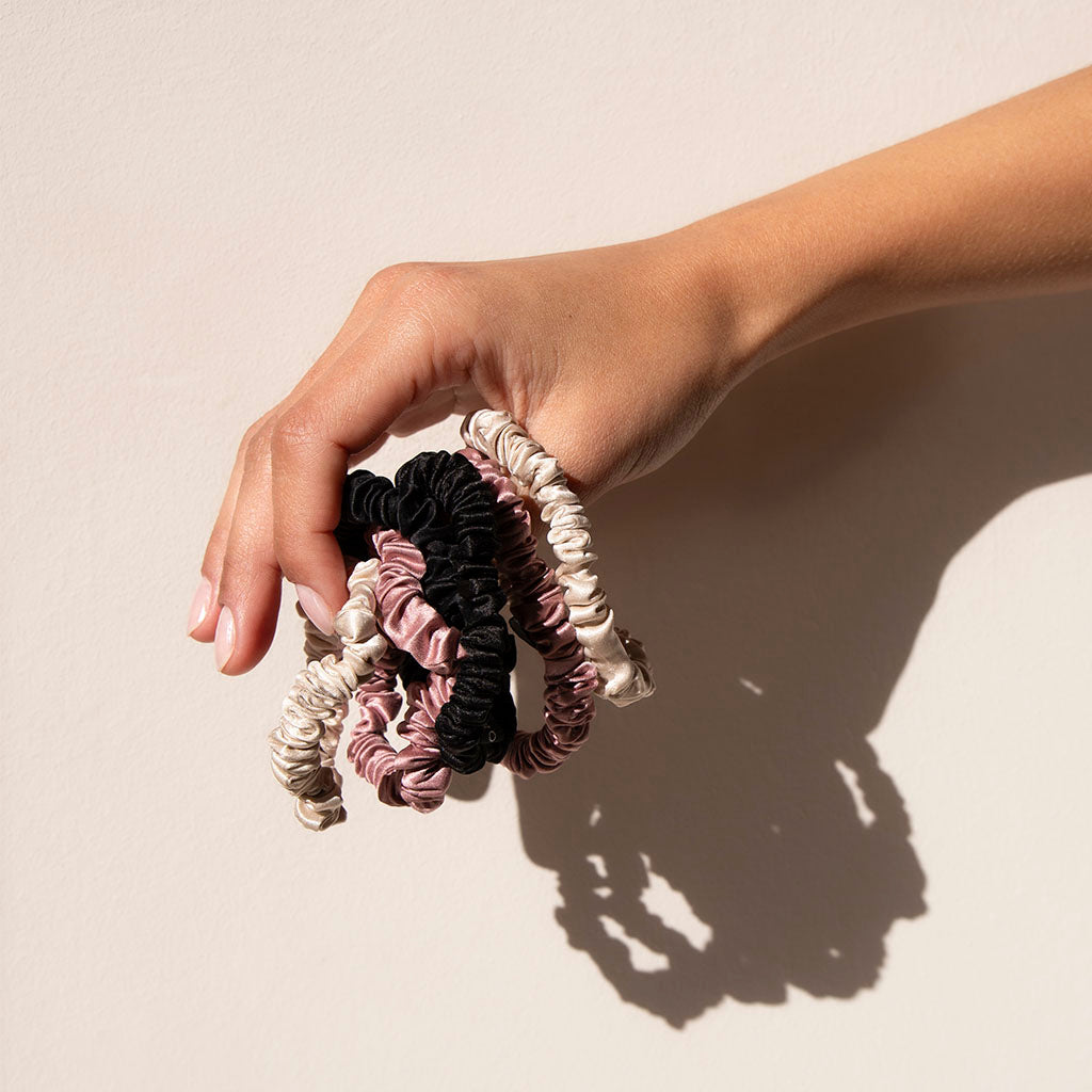 A hand holds the Skinny Scrunchie Set - Rose, Dusty, Black, made from 22 momme silk in pink, beige, and black against a light background. The set casts a soft shadow and provides gentle protection for your hair.
