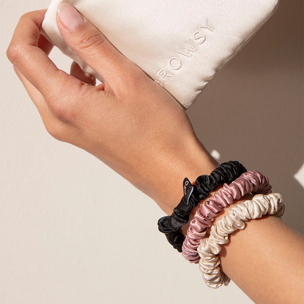 A hand wearing the Skinny Scrunchie Set - Rose, Dusty, Black—made from luxurious 22 momme silk for hair protection—holds a cream pouch embroidered with ROWSY.