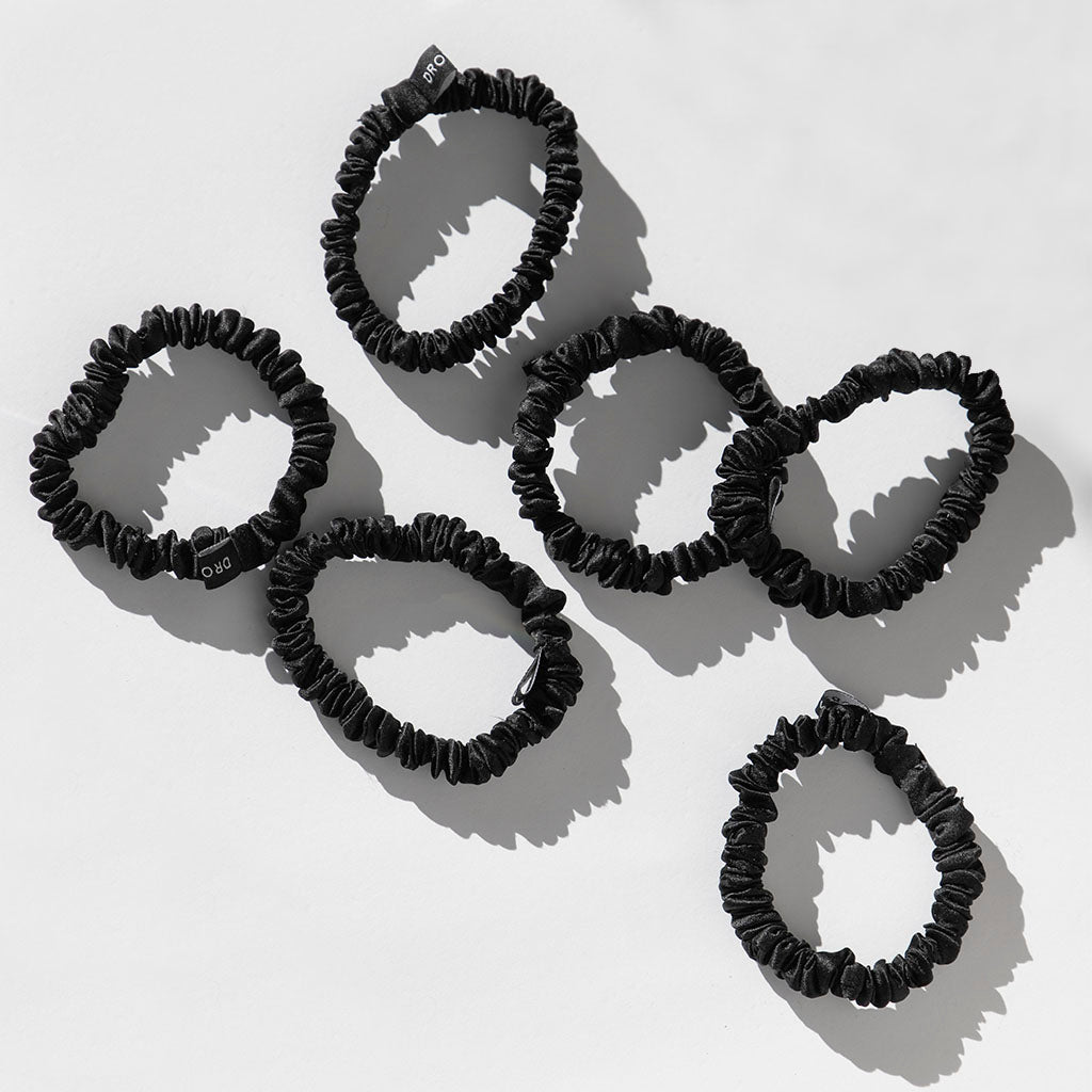 The Skinny Scrunchie Set - Black Jade features six black silk scrunchies with small tags, arranged on a white surface with soft shadows and lighting from the top right.