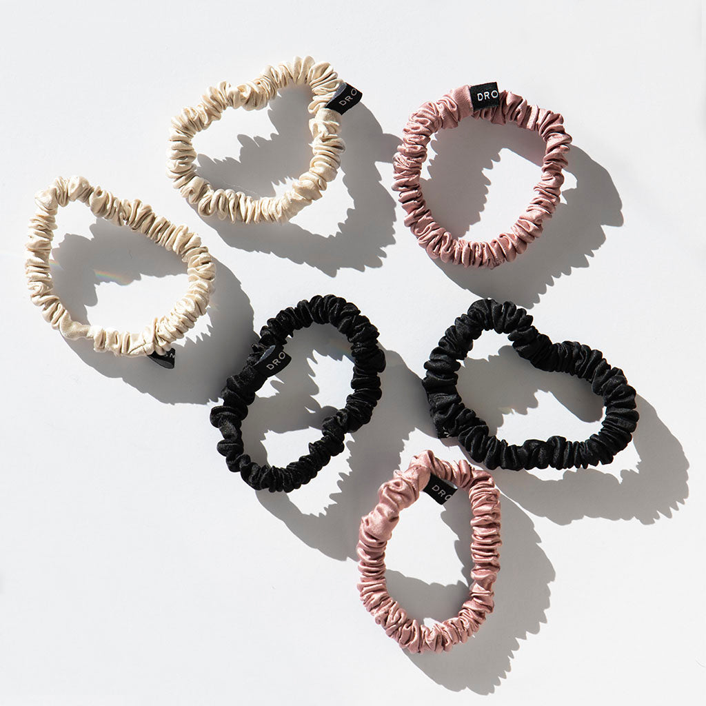 The Skinny Scrunchie Set - Rose, Dusty, Black includes six 22-momme silk scrunchies in cream, black, and pink shades. Each features a small black tag and is arranged in two rows on a white background with soft left shadows.