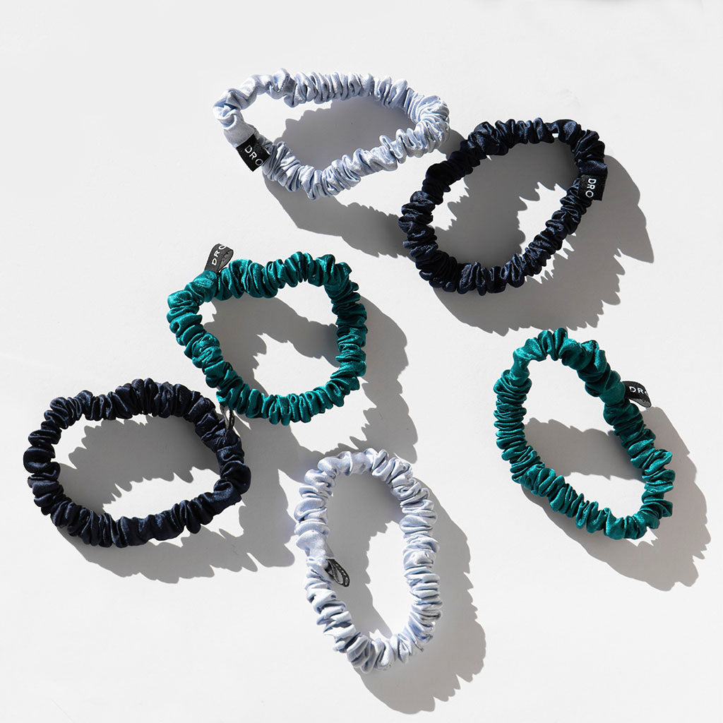 The Skinny Scrunchie Set - Blue & Green Shades includes six silk scrunchies in blue, green, and black tones. Arranged on a white surface, each features a small black tag with white text and offers gentle hair protection.