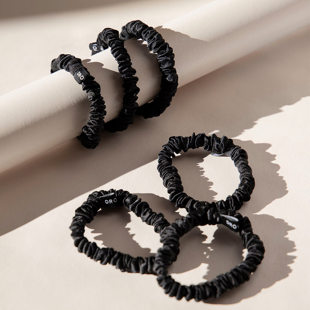 Five black scrunchies from the Skinny Scrunchie Set - Black Jade are shown on a light beige surface; three are stacked on a cylinder and two lay flat. Each scrunchie is made from luxurious 22 momme silk with a soft, gathered texture.
