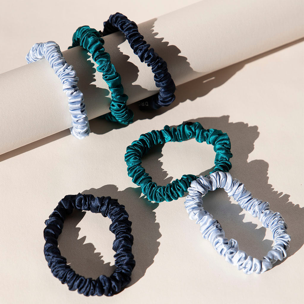 The Skinny Scrunchie Set - Blue & Green Shades features five silk scrunchies in navy, teal, and light blue, shown on a beige surface—some wrapped around a cylinder, others flat—for stylish hair protection.