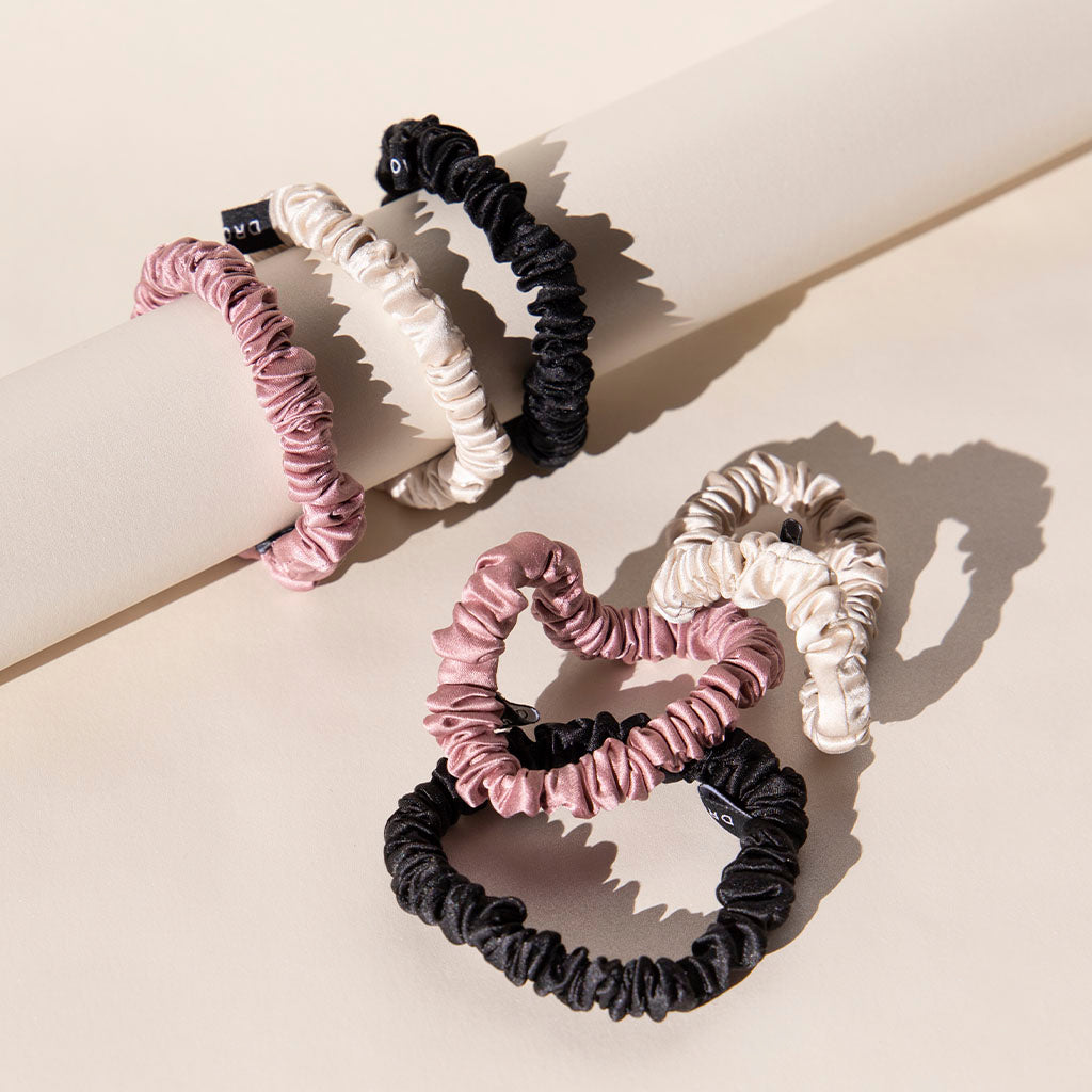 The Skinny Scrunchie Set - Rose, Dusty, Black features three satin scrunchies displayed on and beside a cream cylindrical rod, casting soft shadows while offering gentle hair protection.