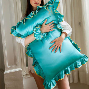A woman indoors holds the drowsysleepco Hollywood Experience bundle - Original: a large turquoise satin pillow with ruffled edges, perfect for beauty sleep. She wears a matching scrunchie on her wrist as natural light filters in.